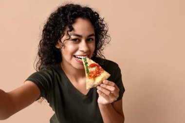 Güzel, genç, mutlu, Afro-Amerikan bir kadın. Lezzetli bir dilim pizzayla bej arka planda selfie çekiyor.