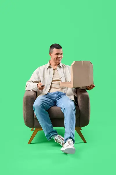 Young man with pizza in armchair watching TV on green background