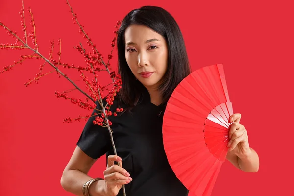 Young Asian woman with fan and berry branches on red background. Chinese New Year celebration