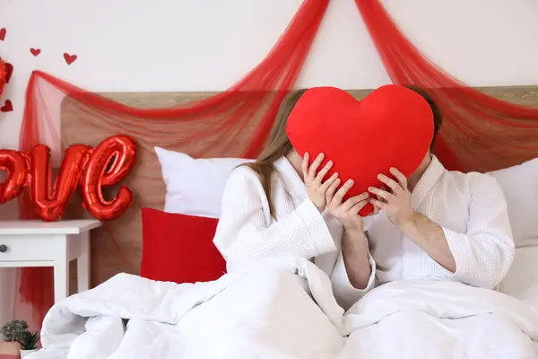 Young couple hiding behind heart-shaped pillow in bedroom on Valentine\'s Day