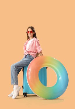 Güneş gözlüklü, bavullu ve turuncu arka planda şişirilebilir yüzüklü küçük kız.