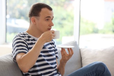 Genç adam evdeki kanepede kahve içiyor.