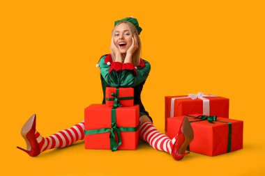 Sarı arka planda Noel hediyesi kutuları olan elf kostümlü duygusal genç bir kadın.
