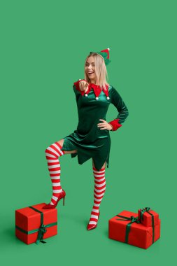 Yeşil arka planda Noel hediyesi kutuları olan elf kostümlü mutlu genç bir kadın.