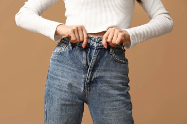 Güzel genç Asyalı kadın kahverengi arka planda şık kot pantolon ilikliyor, yakın plan.