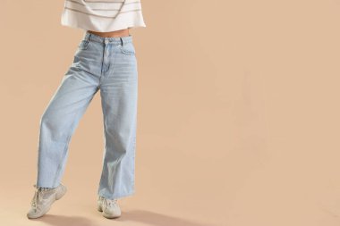 Bej arka planda modaya uygun kot giyen genç bir kadın.