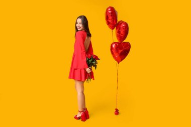 Sarı arka planda kırmızı gül buketi ve kalp şeklinde balonları olan güzel genç bir kadın. Sevgililer Günü kutlaması
