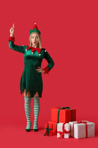 Young woman in elf costume with Christmas gift boxes pointing at something on red background