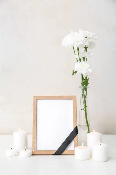 Blank funeral frame, burning candles and vase with flowers on light background