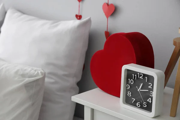 Bedside table with alarm clock and heart-shaped gift box in bedroom, closeup. Valentine\'s Day celebration