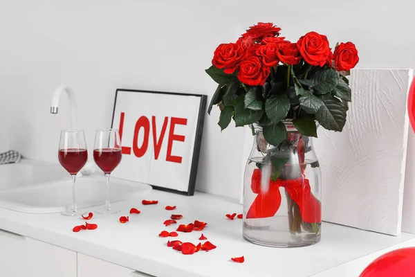 Frame with word LOVE, glasses of wine and red roses on white kitchen counter. Valentine\'s Day celebration