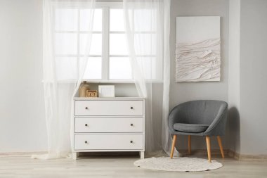 Açık renkli oturma odası, pencerenin yanındaki gri koltuk ve çekmeceler.