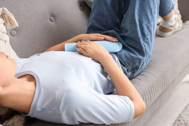 Elinde sıcak su şişesi olan genç bir kadın kanepede yatıyor ve evinde karın ağrısı çekiyor.