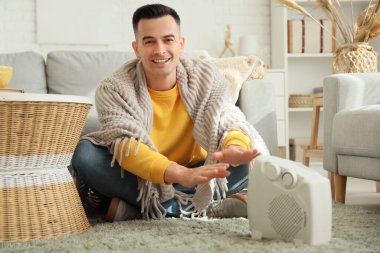 Genç adam elektrikli ısıtıcının yanında ellerini ısıtıyor.