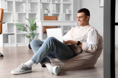 Ofisteki puf minderinde oturan laptoplu yakışıklı genç adam.