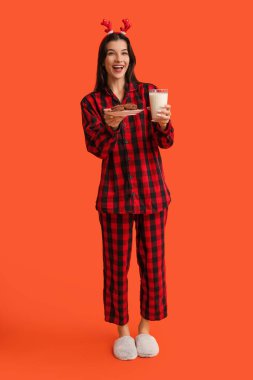 Tatlı kurabiyeler ve turuncu arka planda bir bardak sütle damalı pijama giyen mutlu genç bir kadın.