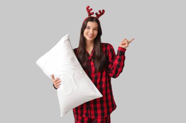 Kareli pijama ve yumuşak yastıklı ren geyiği boynuzları içinde gri arka planda bir şeyi işaret eden güzel genç bir kadın.