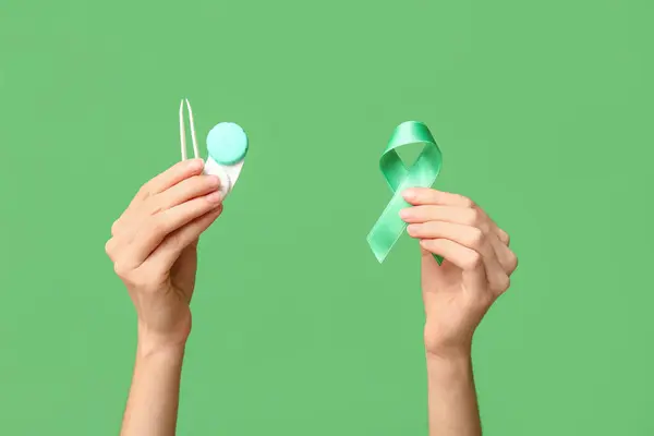 Female hands with ribbon, tweezers and container for contact lenses on green background. Glaucoma awareness concept