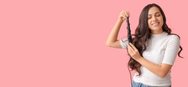 Beautiful young woman using curling iron on pink background with space for text