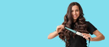 Beautiful young woman with curling iron on blue background with space for text