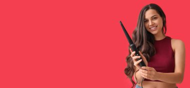 Beautiful young woman with curling iron on red background with space for text