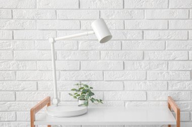 Masa lambası ve çiçek rafta, beyaz tuğla duvarın yanında.