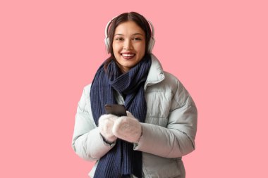 Pembe arka planda cep telefonlu, sıcak fularlı ve kulaklıklı güzel bir Asyalı kadın.