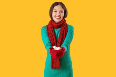 Sarı arka planda marşmelovlu bir fincan kakao tutan sıcak eşarplı güzel bir Asyalı kadın.