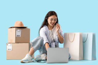 Kulaklıklı genç Asyalı satıcı mavi arka planda dizüstü bilgisayarla çalışıyor.