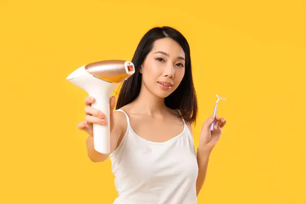Pretty Asian woman with photoepilator and razor on yellow background