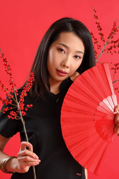 Young Asian woman with fan and berry branches on red background. Chinese New Year celebration