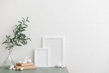 Vase with plant branches, Easter eggs, bunny and blank frames on table near white wall