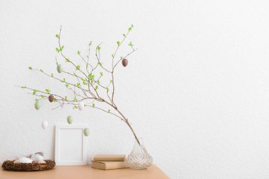 Tree branch with hanging Easter eggs, nest and blank frame on table near white wall