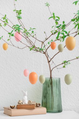 Vase with tree branches, hanging Easter eggs and bunny on table near white wall