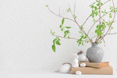 Vase with tree branches, Easter eggs and bunny on table near white wall