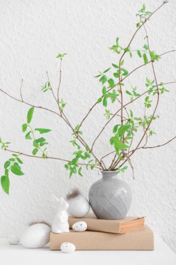 Vase with tree branches, Easter eggs and bunny on table near white wall