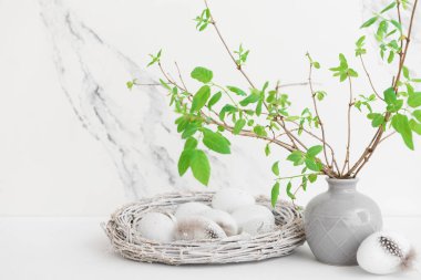 Vase with tree branches and Easter eggs in nest on table near white wall