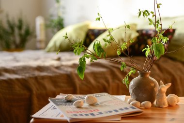 Vase with tree branches, Easter eggs, bunny and magazines on table in interior of bedroom