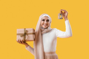 Young Muslim woman with gift and lantern on yellow background. Islamic New Year celebration