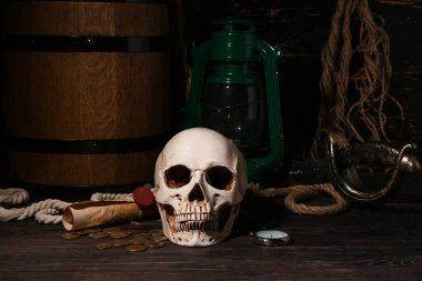 Kılıçlı insan kafatası, cep saati, yağ lambası ve kahverengi ahşap arka planda paralar.