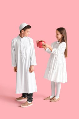 Little girl greeting Muslim boy with gift on pink background