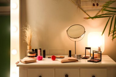 Dressing table with decorative cosmetics, mirror and glowing lamp in dark makeup room