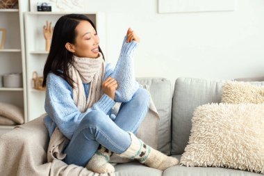 Sıcak kazaklı genç Asyalı kadın evdeki kanepede oturuyor.