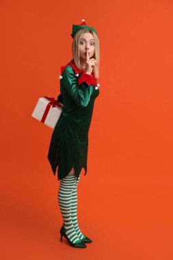 Young woman in elf costume holding Christmas gift box and showing silence gesture on orange background