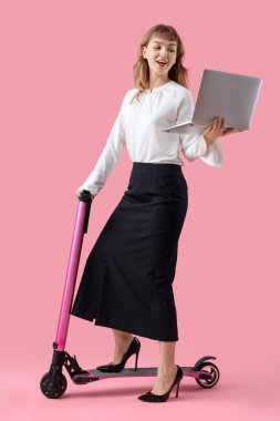 Portrait of young woman with laptop riding kick scooter on pink background