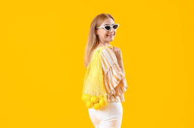 Mature woman holding string bag with lemons on yellow background