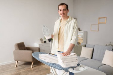 Young man with iron and folded towels on board at home