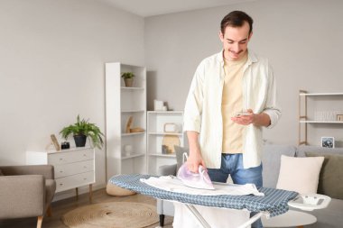 Young man with mobile phone ironing t-shirt on board at home