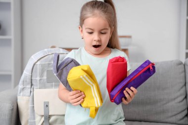 Surprised little schoolgirl with pencil cases at home