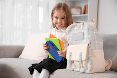 Cute little schoolgirl with copybooks and backpack sitting on sofa at home